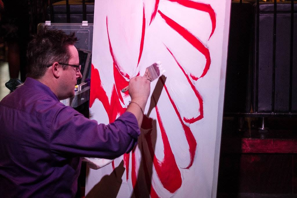 Jim McMahel years ago in a purple shirt, brushing broad red strokes onto a tall white canvas at an event